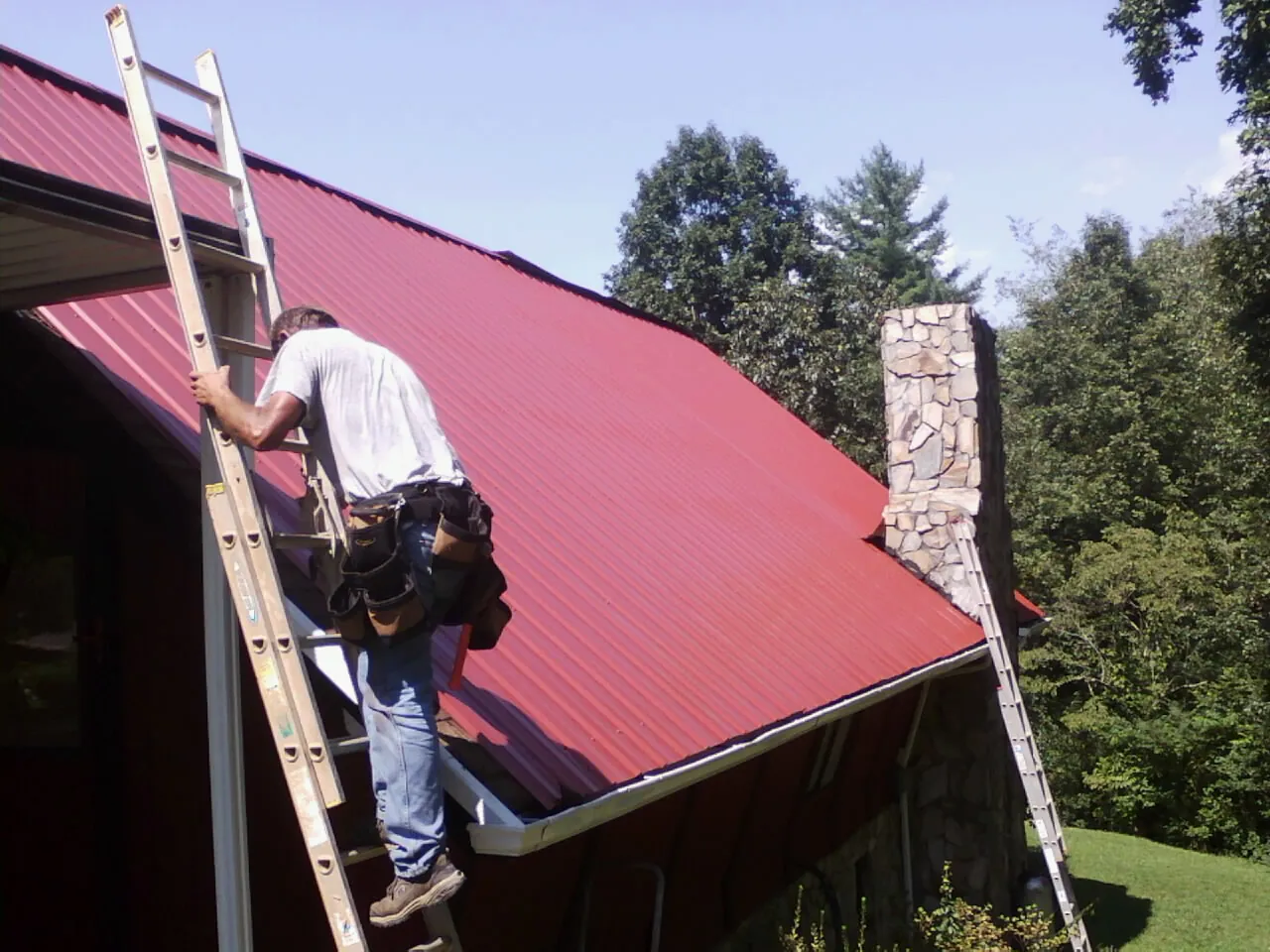 Roofer on a ladder beside a red metal roof and stone chimney — Soffit & Fascia Repair by Roofing Brothers in Grassy Creek